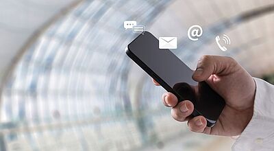 Eine Hand hält ein Smartphone vor einem modernen, verglasten Hintergrund. Über dem Gerät schweben Symbole für Kommunikation: eine Sprechblase, ein Briefumschlag, ein @-Zeichen und ein Telefonhörer – sie symbolisieren Messaging, E-Mail, Internet und Telefonie.