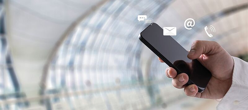 Eine Hand hält ein Smartphone vor einem modernen, verglasten Hintergrund. Über dem Gerät schweben Symbole für Kommunikation: eine Sprechblase, ein Briefumschlag, ein @-Zeichen und ein Telefonhörer – sie symbolisieren Messaging, E-Mail, Internet und Telefonie.