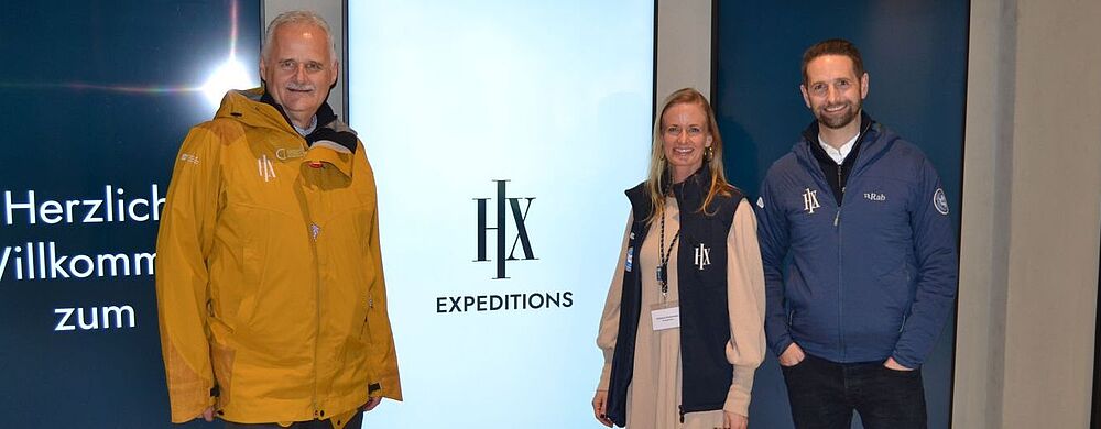 Zwei Männer und eine Frau – Führungsrege einer Reederei – posiert vor einem Display mit dem Firmenlogo „HX“.