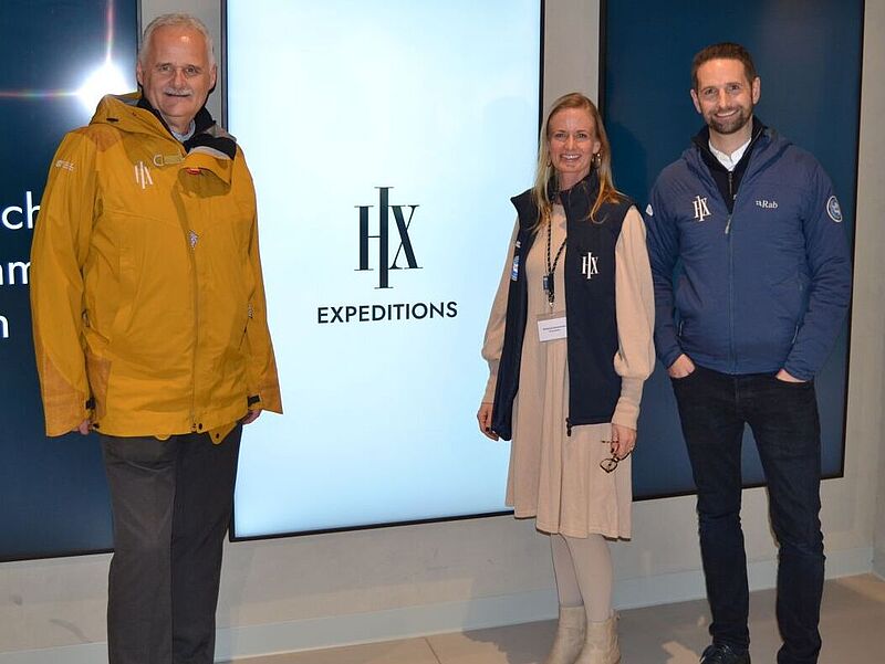 CEO Gebhard Rainer, Sales Director Kimberly Setzermann und Europa-Chef Nathaniel Sherborne beim Partner-Event in Hamburg. Foto: ck Zwei Männer und eine Frau – Führungsrege einer Reederei – posiert vor einem Display mit dem Firmenlogo „HX“.