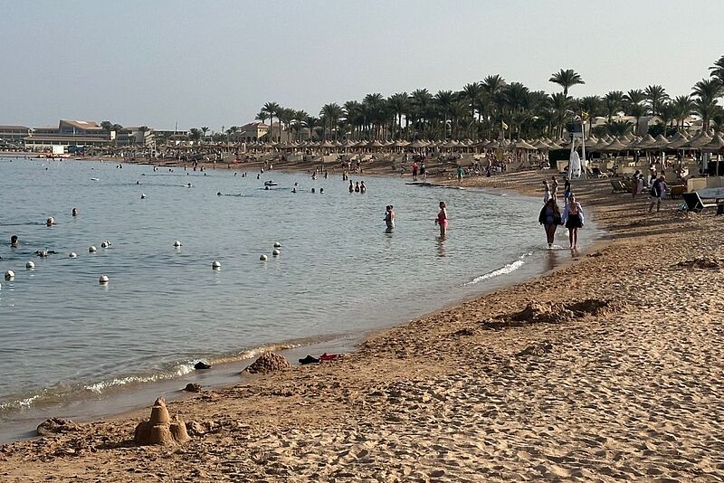 Das Bild zeigt einen Strand in Ägypten, Spaziergänger laufen an der Wasserlinie entlang, Schwimmer im Meer und Liegen an den Stränden. Im Hintergrund eine Hotelanlage.