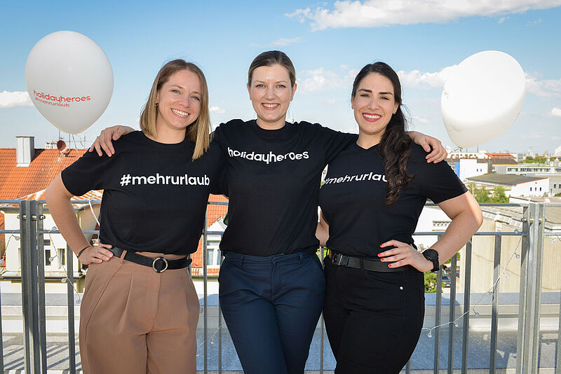 Drei lächelnde Frauen stehen nebeneinander auf einer sonnigen Dachterrasse mit Blick auf ein städtisches Umfeld. Sie tragen schwarze T-Shirts mit weißen Aufdrucken – zwei mit dem Hashtag „#mehrurlaub“ und eines mit „holidayheroes“. Zwei weiße Luftballons mit der Aufschrift „holidayheroes #mehrurlaub“ schweben im Hintergrund. Alle Frauen wirken freundlich und verbunden, mit Armen umeinander gelegt. Der Himmel ist blau mit einigen Wolken.