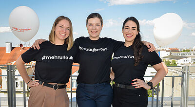 Drei lächelnde Frauen stehen nebeneinander auf einer sonnigen Dachterrasse mit Blick auf ein städtisches Umfeld. Sie tragen schwarze T-Shirts mit weißen Aufdrucken – zwei mit dem Hashtag „#mehrurlaub“ und eines mit „holidayheroes“. Zwei weiße Luftballons mit der Aufschrift „holidayheroes #mehrurlaub“ schweben im Hintergrund. Alle Frauen wirken freundlich und verbunden, mit Armen umeinander gelegt. Der Himmel ist blau mit einigen Wolken.
