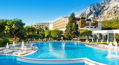 Für Alleinreisende bietet TUI nun Solowochen an, unter anderem im November im TUI Magic Life Club Beldibi in der Türkei Hotelanlage im Grünen mit Bergen im Hintergrund und zwei Pools im Vordergrund. Der Pool ist umgeben von weißen Liegen und Sonnenschirmen. Der Himmel ist blau.
