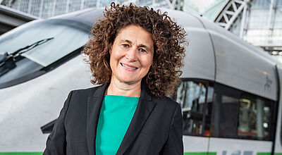 Eine Managerin mittleren Alters mit dunklem Sakko und grüner Bluse steht lächelnd vor einem ICE-Zug vor einem Bahnhofsgebäude.