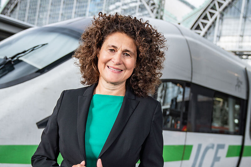 Eine Managerin mittleren Alters mit dunklem Sakko und grüner Bluse steht lächelnd vor einem ICE-Zug vor einem Bahnhofsgebäude.