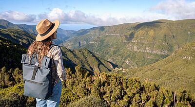 Das Foto zeigt eine Frau mit einem Wanderrucksack auf dem Rücken von hinten, die über eine grüne Hügellandschaft blickt.