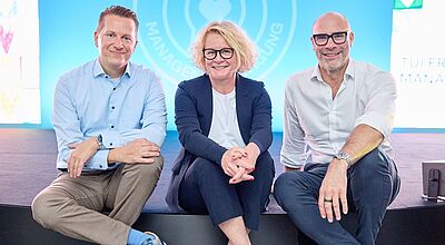 Drei Personen sitzen lächelnd nebeneinander auf einer Bühne vor einem blauen Hintergrund mit dem Logo „TUI“. Die Frau in der Mitte trägt einen dunkelblauen Hosenanzug und rosa Sneaker, die beiden Männer links und rechts daneben sind leger in Hemd und Hose gekleidet und tragen ebenfalls sportliche Schuhe.