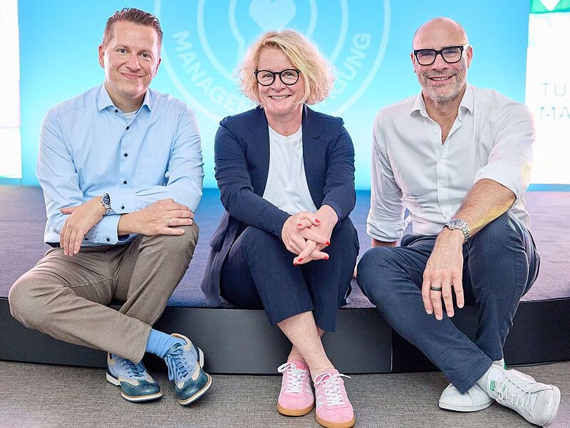 Drei Personen sitzen lächelnd nebeneinander auf einer Bühne vor einem blauen Hintergrund mit dem Logo „TUI“. Die Frau in der Mitte trägt einen dunkelblauen Hosenanzug und rosa Sneaker, die beiden Männer links und rechts daneben sind leger in Hemd und Hose gekleidet und tragen ebenfalls sportliche Schuhe.