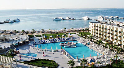 Blick aus erhöhter Position über eine Hotelanlage mit Pool, im Hintergrund das Meer und eine Marina, an der mehrere Boote liegen