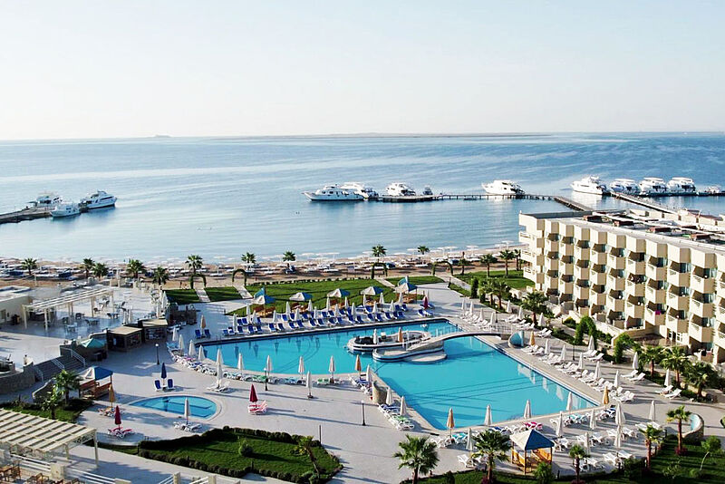 Blick aus erhöhter Position über eine Hotelanlage mit Pool, im Hintergrund das Meer und eine Marina, an der mehrere Boote liegen
