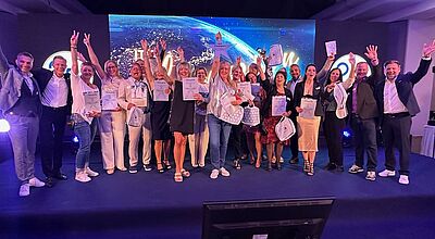 Eine Gruppe von etwa 25 fröhlichen Menschen steht auf einer Bühne und hält Zertifikate, Taschen und Auszeichnungen in die Luft. Die Personen jubeln, lachen und posieren gemeinsam vor einem großen Bildschirm im Hintergrund, der eine Weltkarte mit Lichtern und den Schriftzug „Champion“ zeigt. Die Atmosphäre ist festlich und feierlich. Es ist die Preisverleihung der Reisebüro-Kooperation TUI Travel Star.