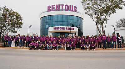 Gruppenfoto eines großen Teams vor einem modernen, verglasten Firmengebäude mit der Aufschrift 'BENTOUR REISEN'. Die Mitarbeitenden tragen einheitliche lila Oberteile oder Jacken und stehen bzw. sitzen in einer langen Reihe auf einer Straße vor dem Gebäude. Der Himmel ist bewölkt, und es gibt Bäume sowie Straßenlaternen im Hintergrund.