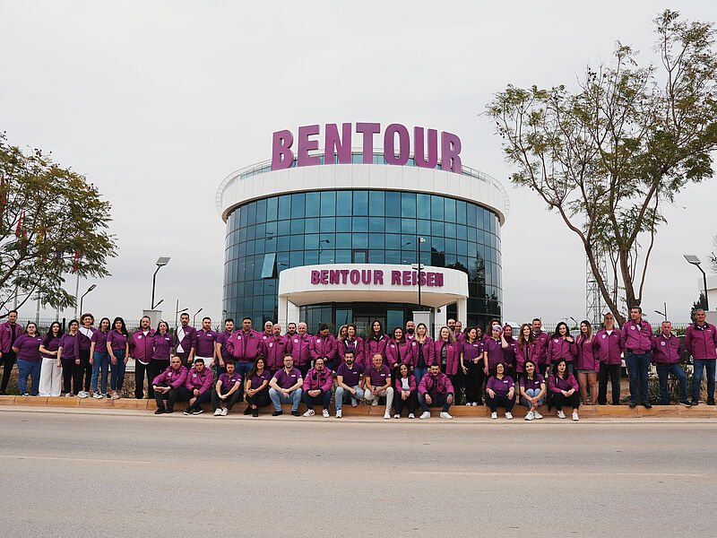 Gruppenfoto eines großen Teams vor einem modernen, verglasten Firmengebäude mit der Aufschrift 'BENTOUR REISEN'. Die Mitarbeitenden tragen einheitliche lila Oberteile oder Jacken und stehen bzw. sitzen in einer langen Reihe auf einer Straße vor dem Gebäude. Der Himmel ist bewölkt, und es gibt Bäume sowie Straßenlaternen im Hintergrund.