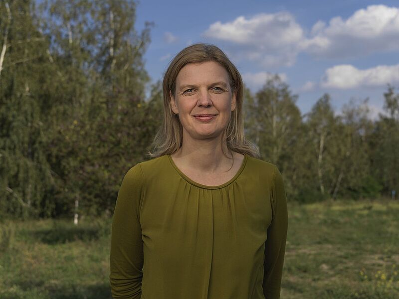 Antje Monshausen, Geschäftsführerin von Ecpat Deutschland e.V., steht bei Tageslicht vor einer naturbelassenen Kulisse mit Bäumen und Wiese. Sie trägt eine olivgrüne Bluse und blickt freundlich in die Kamera. Der Himmel ist leicht bewölkt.
