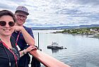 Ein Mann und eine Frau mit Brillen lehnen an der Reling, rechts ist der Oslo-Fjord zu sehen mit Festland und einem kleinen Schiff auf dem Wasser.