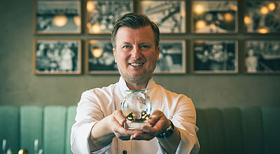 Sternekoch Kevin Fehling in weißer Uniform präsentiert eine Kristalltrophäe, im Hintergrund sind alte Fotografien und eine grüne Restauranteinrichtung zu erkennen.