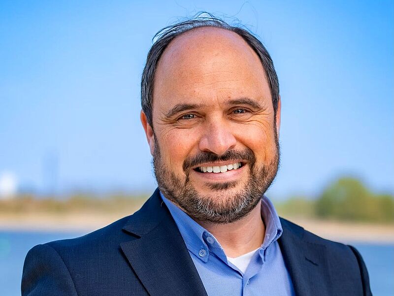 Hurtigruten-Manager Marius Griego hat die Reederei auf den Reisebüro-Vertrieb ausgerichtet. Ein Reederei-Manager mit dunkelbraunem Haar, hoher Stirn und Bart blickt lächelnd in die Kamera. Er trägt ein dunkles Sakko auf blauem Oberhemd, im Hintergrund ist Wasser und ein Flussufer zu sehen.