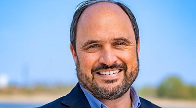 Ein Reederei-Manager mit dunkelbraunem Haar, hoher Stirn und Bart blickt lächelnd in die Kamera. Er trägt ein dunkles Sakko auf blauem Oberhemd, im Hintergrund ist Wasser und ein Flussufer zu sehen. 