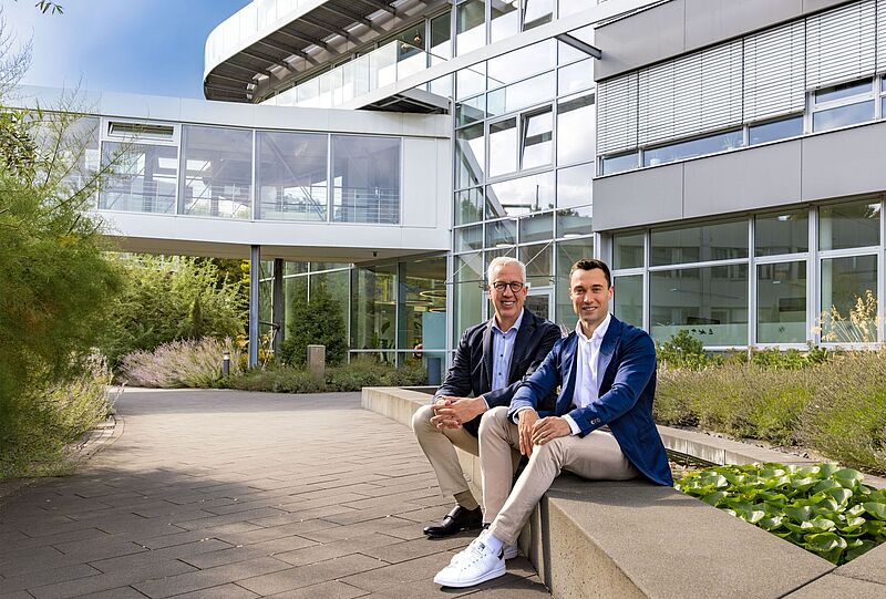 Zwei Geschäftsmänner in Business Casual sitzen entspannt auf einer Mauer vor einem modernen Bürogebäude mit Glasfassade und Gartenbepflanzung.