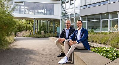 Gerald (links) und Steffen Kassner vor der Unternehmenszentrale in Duisburg Zwei Geschäftsmänner in Business Casual sitzen entspannt auf einer Mauer vor einem modernen Bürogebäude mit Glasfassade und Gartenbepflanzung.