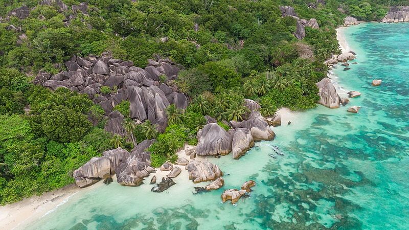 Luftaufnahme eines Seychellen-Strandes mit markanten grauen Granitfelsen, türkisfarbenem Wasser und tropischer Vegetation.