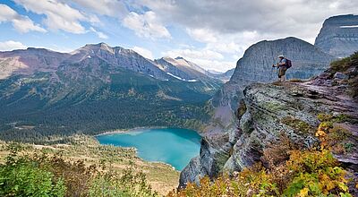 Im Glacier Nationalpark blickt eine Frau von einem steilen Felsvorsprung auf einen türkisblauen Bergsee und die beeindruckende Bergkulisse in Montana, USA. Die Natur ist üppig, mit buntem Herbstlaub im Vordergrund und weiten Nadelwäldern im Tal. 