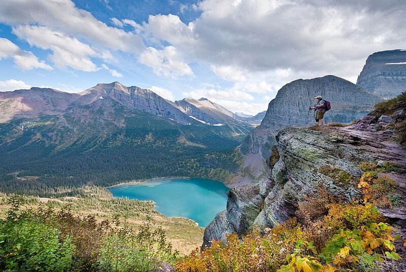 Im Glacier Nationalpark blickt eine Frau von einem steilen Felsvorsprung auf einen türkisblauen Bergsee und die beeindruckende Bergkulisse in Montana, USA. Die Natur ist üppig, mit buntem Herbstlaub im Vordergrund und weiten Nadelwäldern im Tal. 
