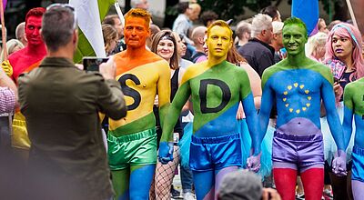 Feiernde beim CSD 2024 in Frankfurt am Main. In diesem Jahr feiert auch die Touristik mit Eine Gruppe von Menschen nimmt an einer LGBTQ+-Parade teil. Im Vordergrund stehen fünf Männer mit bunt bemalten Oberkörpern und passenden kurzen Hosen und Tüllröcken in Regenbogenfarben. Zwei von ihnen haben große schwarze Buchstaben „S“ und „D“ auf die Brust gemalt, der dritte zeigt gelbe Sterne in Kreisform wie die EU-Flagge. Sie halten sich an den Händen und lächeln. Im Hintergrund fotografieren Zuschauer die Szene, während weitere Teilnehmende mit Regenbogen- und Pride-Flaggen zu sehen sind.