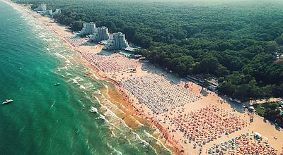 Luftaufnahme eines Strandabschnitts mit türkisfarbenem Meer, Wellenbrechern, weißen Hotelgebäuden und symmetrisch angeordneten Sonnenschirmen am Waldrand.
