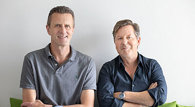 Stolz auf über eine Million Buchungen: die Sunny-Cars-Chefs Kai Sannwald und Thorsten Lehmann. Foto: Sunny Cars Zwei lächelnde Männer mittleren Alters in legerer Kleidung sitzen nebeneinander auf einem grünen Sofa vor einer grau-weißen Wand