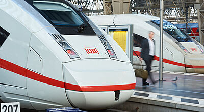 Ein Geschäftsreisender mit Bewegungsunschärfe geht in einer Bahnhofshalle auf einem Bahnsteig und wird eingerahmt von zwei weiß-roten ICE-Zügen