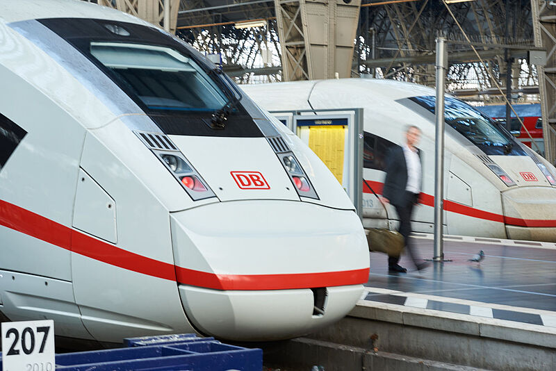 Ein Geschäftsreisender mit Bewegungsunschärfe geht in einer Bahnhofshalle auf einem Bahnsteig und wird eingerahmt von zwei weiß-roten ICE-Zügen