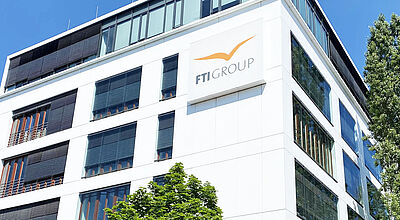 Zentrale von FTI in München: Zurzeit sind noch 40 Beschäftigte vor Ort Moderne Bürogebäude-Fassade mit dem Logo der FTI GROUP an der Außenwand. Der Himmel ist klar und blau, grüne Bäume sind im Vordergrund zu sehen. Das Gebäude hat große Fenster mit dunklen Jalousien und eine helle Fassade.