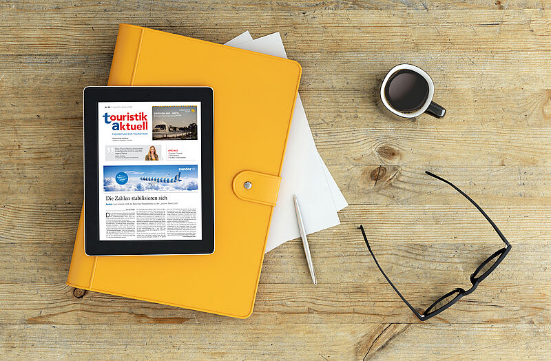 Ein Tablet mit der geöffneten Online-Ausgabe des Magazins 'touristik aktuell' liegt auf einer gelben Mappe auf einem Holztisch. Daneben befinden sich lose Blätter, ein silberner Kugelschreiber, eine schwarze Brille und eine Tasse schwarzer Kaffee.