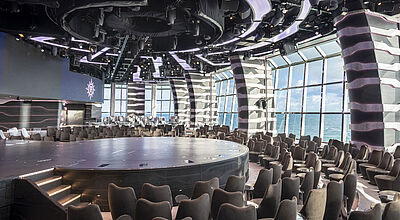 In der Carousel Lounge werden ab diesem Jahr zwei neue Cirque-Aerial-Shows gezeigt – eine ab Juni und die andere ab dem Herbst Blick in die Entertainment-Lounge eines Kreuzfahrtschiffes. Um die leere Rundbühne sind Sessel angeordnet, oberhalb der Bühne hängt Technik an der Decke. Hinter den Zuschauerplätzen ist durch große Fenster das Meer zu sehen.