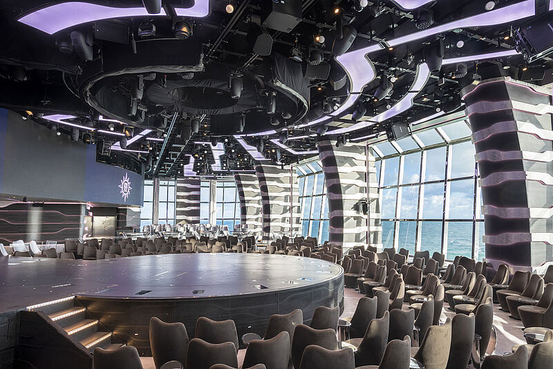 In der Carousel Lounge werden ab diesem Jahr zwei neue Cirque-Aerial-Shows gezeigt – eine ab Juni und die andere ab dem Herbst Blick in die Entertainment-Lounge eines Kreuzfahrtschiffes. Um die leere Rundbühne sind Sessel angeordnet, oberhalb der Bühne hängt Technik an der Decke. Hinter den Zuschauerplätzen ist durch große Fenster das Meer zu sehen.