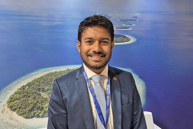 Ibrahim Shiuree, CEO der Maldives Marketing Corporation, peilt für dieses Jahr 2,3 Millionen Besucher an Freundlicher junger Mann im Anzug vor einer Fotowand mit kleinen Inseln im türkisgrünen Meer