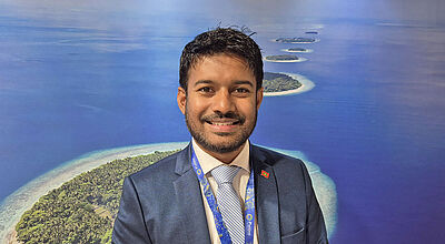 Freundlicher junger Mann im Anzug vor einer Fotowand mit kleinen Inseln im türkisgrünen Meer