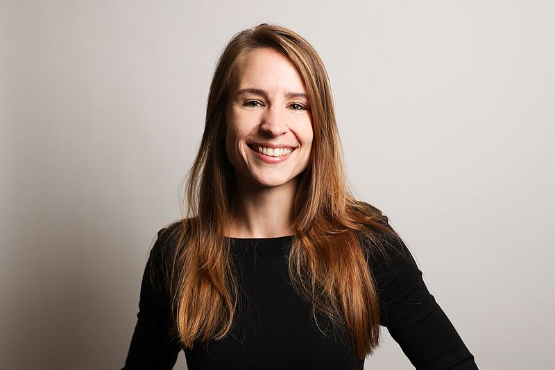 Eine junge Frau mit langen, hellbraunen Haaren lächelt in die Kamera. Sie trägt ein schwarzes Oberteil und steht vor einem hellen, neutralen Hintergrund.