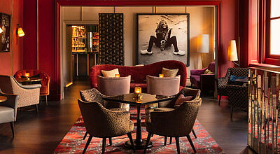 Elegantes Interieur einer Bar mit roten Wänden, gemütlichen Sitzgruppen und großem Schwarz-Weiß-Foto einer Musikerin mit Saxophon an der Wand.