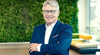 Ein Mann in dunkelblauem Anzug mit weißem Hemd und Brille steht mit verschränkten Armen lächelnd vor einer begrünten Wand in einem modernen Bürogebäude.