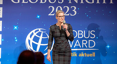 Fokus auf die Reisebüros: Jeanette Buller 2023 auf der Bühne des Globus Award von touristik aktuell. Foto: ta Eine Frau in einem karierten, knielangen Kleid steht mit einem Mikrofon in der Hand auf einer Bühne vor einer blau beleuchteten Wand mit der Aufschrift „Globus Night 2023“ und „Globus Award“, während im Vordergrund unscharf das Publikum zu sehen ist.