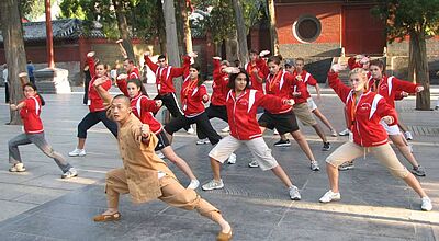 Gruppe junger Menschen in roten Jacken übt gemeinsam mit einem Shaolin-Mönch traditionelle Kung-Fu-Bewegungen im Innenhof eines Tempels in China.