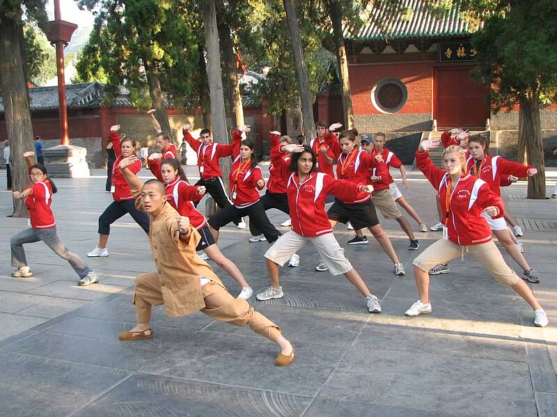 Gruppe junger Menschen in roten Jacken übt gemeinsam mit einem Shaolin-Mönch traditionelle Kung-Fu-Bewegungen im Innenhof eines Tempels in China.