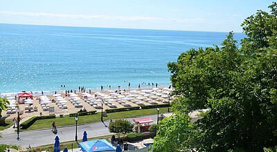 Bulgarisches Topziel: Goldstrand bei Varna. Blick von oben auf einen sonnigen Sandstrand mit vielen weißen Sonnenliegen und Sonnenschirmen, badenden Menschen im türkisblauen Meer und einer Promenade mit Bäumen und kleinen Verkaufsständen im Vordergrund.