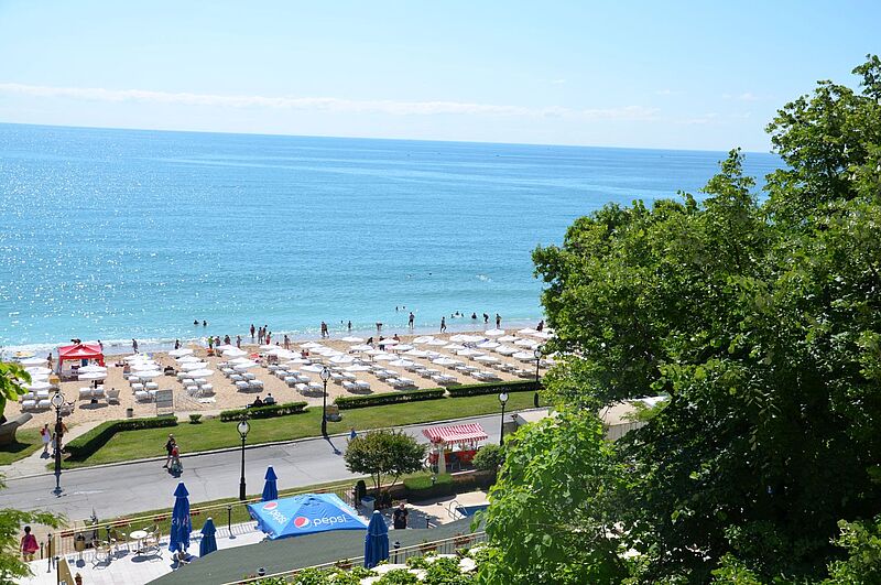 Bulgarisches Topziel: Goldstrand bei Varna. Blick von oben auf einen sonnigen Sandstrand mit vielen weißen Sonnenliegen und Sonnenschirmen, badenden Menschen im türkisblauen Meer und einer Promenade mit Bäumen und kleinen Verkaufsständen im Vordergrund.