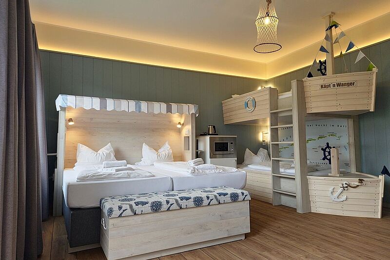 Gemütlich eingerichtetes Hotelzimmer mit maritimem Thema. Ein großes Doppelbett mit einem Strandkorb-ähnlichen Kopfteil steht im Vordergrund, daneben eine Truhe mit maritimen Motiven. Im Hintergrund befinden sich kreative Etagenbetten in Schiffsform mit einer Leiter, kleinen Segeln und nautischen Details. Die Wand ist in einem ruhigen Blau gehalten, und indirekte Beleuchtung schafft eine warme Atmosphäre. Ein Netzlampenschirm an der Decke verstärkt das maritime Ambiente