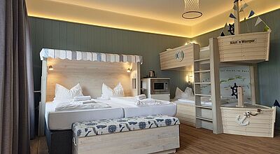 Gemütlich eingerichtetes Hotelzimmer mit maritimem Thema. Ein großes Doppelbett mit einem Strandkorb-ähnlichen Kopfteil steht im Vordergrund, daneben eine Truhe mit maritimen Motiven. Im Hintergrund befinden sich kreative Etagenbetten in Schiffsform mit einer Leiter, kleinen Segeln und nautischen Details. Die Wand ist in einem ruhigen Blau gehalten, und indirekte Beleuchtung schafft eine warme Atmosphäre. Ein Netzlampenschirm an der Decke verstärkt das maritime Ambiente