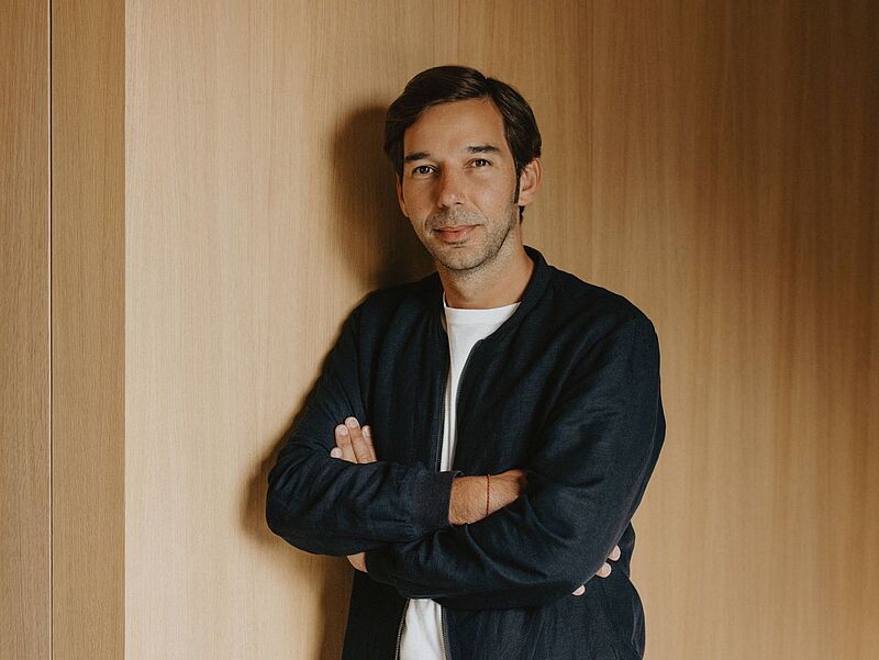 Ein Mann mittleren Alters steht vor einer holzvertäfelten Wand, mit verschränkten Armen und leichtem Lächeln, gekleidet in einer dunklen Jacke und einem weißen T-Shirt.